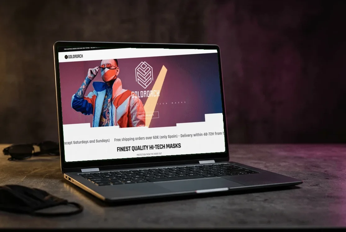 La portada de la tienda online de Goldroach en la pantalla de un portátil, sobre una mesa con una mascarilla negra y unas gafas de sol.